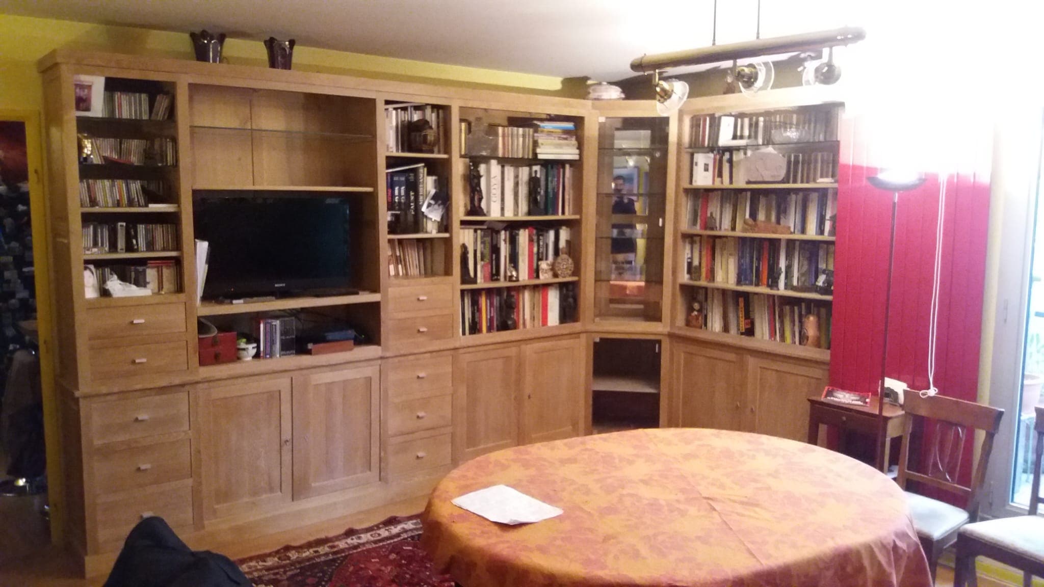 Agencement de bibliothèque en chêne massif toute hauteur tournant l’angle d’un salon : rayonnages ouverts, colonnes de tiroirs, portes à panneaux, niche télévision et vitrine d’angle