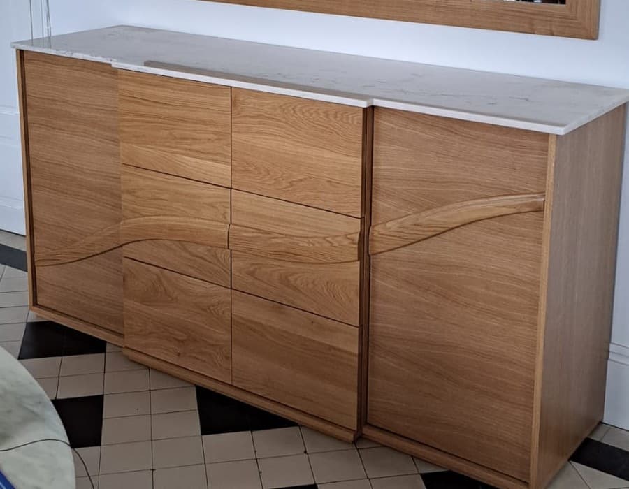 Buffet contemporain en chêne massif à plateau de marbre blanc veiné : deux portes encadrant une colonne de tiroirs, façade traversée par une vague sculptée servant de prise de main