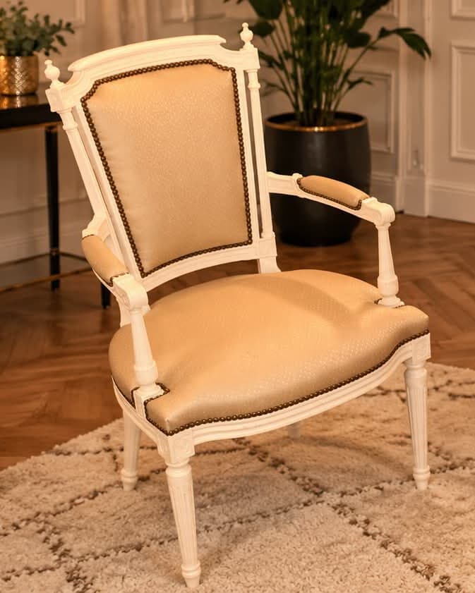 Fauteuil Louis XVI laqué blanc à dossier à chapeau, colonnettes cannelées surmontées de toupies, garni d’un tissu champagne bordé de clous de laiton, manchettes garnies et pieds fuselés cannelés